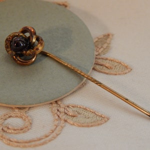 May include: A gold-toned pin with a knotted design and a dark purple stone in the center. The pin is on a light gray surface.