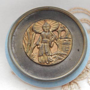 May include: A circular button featuring a central gold-toned metal inlay depicting a figure holding an umbrella, surrounded by a dark metallic ring and a light blue outer border. The design includes raised details of bamboo and a small structure.