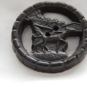 May include: A black plastic button with a raised design of a person riding a horse. The button has a textured rim and two holes in the center.