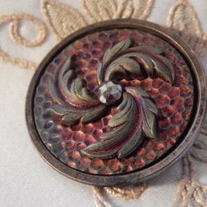 May include: A vintage button with a raised design of swirling leaves in shades of green and brown. The button has a textured, iridescent background in shades of red and copper. A clear, faceted glass stone is set in the center of the button.