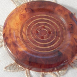 Puede incluir: Un botón grande, redondo y marrón con un diseño de círculos concéntricos. El botón está hecho de un material translúcido con un patrón de mármol.