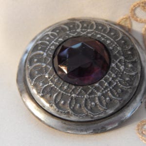 May include: A circular silver-toned metal button featuring an intricate embossed pattern of curved lines and dots surrounding a central, faceted, deep purple gemstone. The button has a smooth, raised outer rim and a dark, weathered metallic finish.