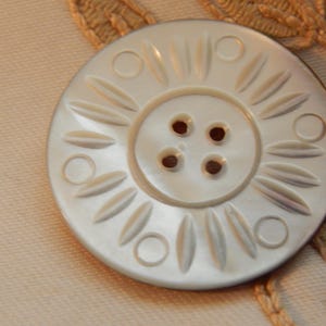 May include: A large, round, off-white button with a decorative floral design. The button has four holes in the center and is surrounded by a pattern of elongated ovals and circles. The button is set against a light beige fabric.