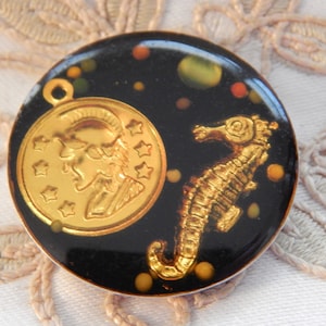 May include: A round black button with a gold-colored seahorse and a gold-colored medallion with a profile of a person wearing a helmet. The button has a black background with gold-colored dots.