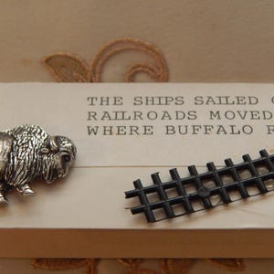 Könnte beinhalten: Ein silberner Büffel-Ansteckpin und ein kleines schwarzes Kunststoff-Gleisstück liegen auf einer cremefarbenen Oberfläche. Der Text "THE SHIPS SAILED ONWA...RAILROADS MOVED WES...WHERE BUFFALO ROAM..." ist darüber gedruckt.