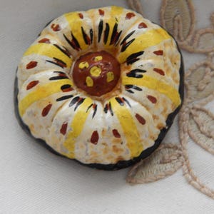 Op de afbeelding: Handbeschilderde decoratieve keramische knoop met een uniek ontwerp. De knoop heeft een centrale bruine cirkel omgeven door witte, gele en zwarte gestreepte secties, met rode spikkels. De knoop heeft een zwarte rand.