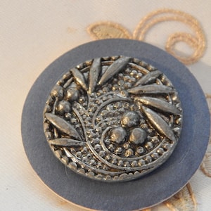 May include: A detailed, antique silver button with an intricate floral design, including leaves and small round accents. The button is set on a circular, slate-blue backing, showcasing its ornate details. The button is a vintage item.