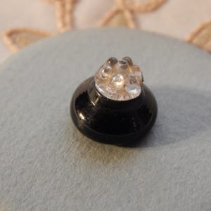 May include: A small, black button with a clear, textured top. The button is round and has a glossy finish. It is set on a light blue felt-like surface, with a hint of a floral design in the background.