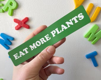 May include: A green rectangular magnet with the words "EAT MORE PLANTS" in white lettering. The magnet is held by a hand, with colorful magnetic letters scattered around it.
