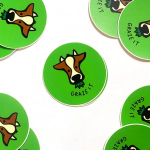 May include: Green circular stickers with a cartoon cow face in brown and white, eating grass. The text "GRAZE IT" is below the cow's face in black. The stickers are scattered on a white surface.