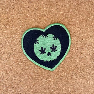 Puede incluir: Un parche en forma de corazón con un contorno negro y un borde verde. Dentro del corazón hay un diseño de calavera negra con hojas de cannabis verdes para los ojos y la boca. El fondo es una superficie texturizada y marrón claro.