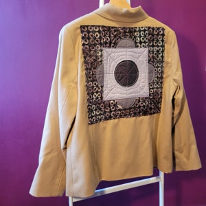 May include: A tan blazer with a decorative quilted patch on the back. The patch features a geometric design with shades of brown, gray, and black. The blazer is hanging on a wooden hanger.