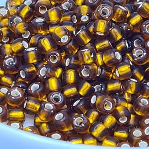 Pode incluir: Um close-up de um monte de pequenas contas redondas de vidro marrom e dourado. As contas t&ecirc;m um padr&atilde;o listrado e um furo prateado no centro.