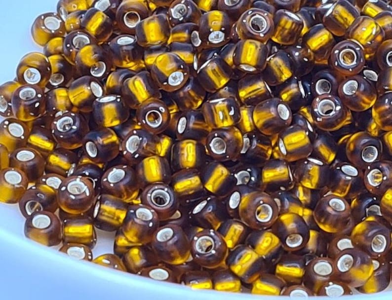 Pode incluir: Um close-up de um monte de pequenas contas redondas de vidro marrom e dourado. As contas t&ecirc;m um padr&atilde;o listrado e um furo prateado no centro.