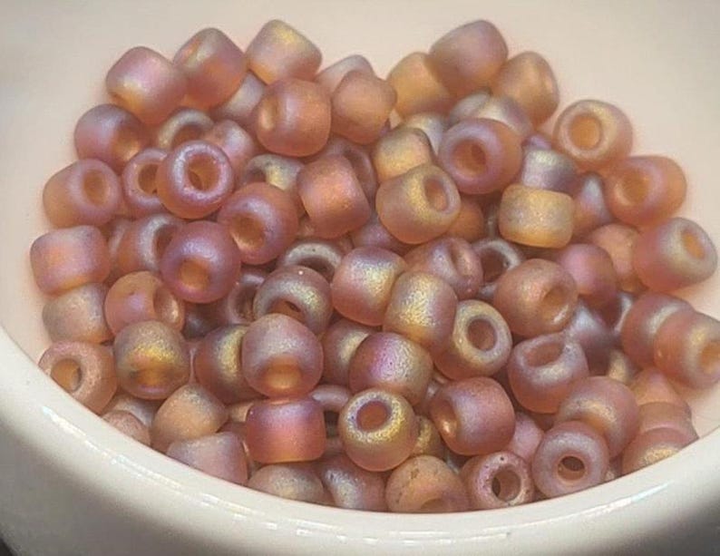 Pode incluir: Um monte de pequenas contas de vidro redondas e iridescentes de cor rosa claro. As contas t&ecirc;m um brilho ligeiramente met&aacute;lico e s&atilde;o perfeitas para a fabrica&ccedil;&atilde;o de joias.