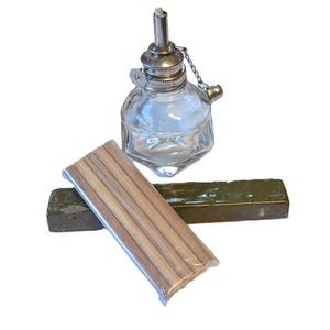 Puede incluir: Una lámpara de aceite de vidrio con una parte superior plateada y una cadena. La lámpara está sobre una barra de jabón verde y junto a un paquete de mechas de madera.