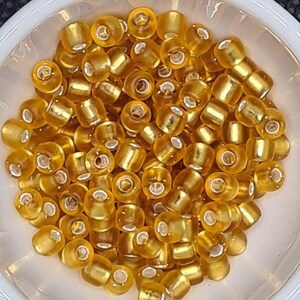 Pode incluir: Uma tigela de pequenas contas de vidro redondas de cor dourada. As contas t&ecirc;m um brilho met&aacute;lico e est&atilde;o dispostas em um padr&atilde;o aleat&oacute;rio.
