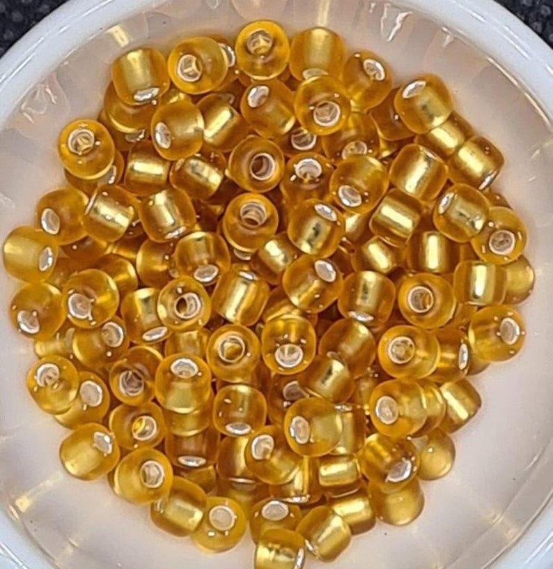 Pode incluir: Uma tigela de pequenas contas de vidro redondas de cor dourada. As contas t&ecirc;m um brilho met&aacute;lico e est&atilde;o dispostas em um padr&atilde;o aleat&oacute;rio.