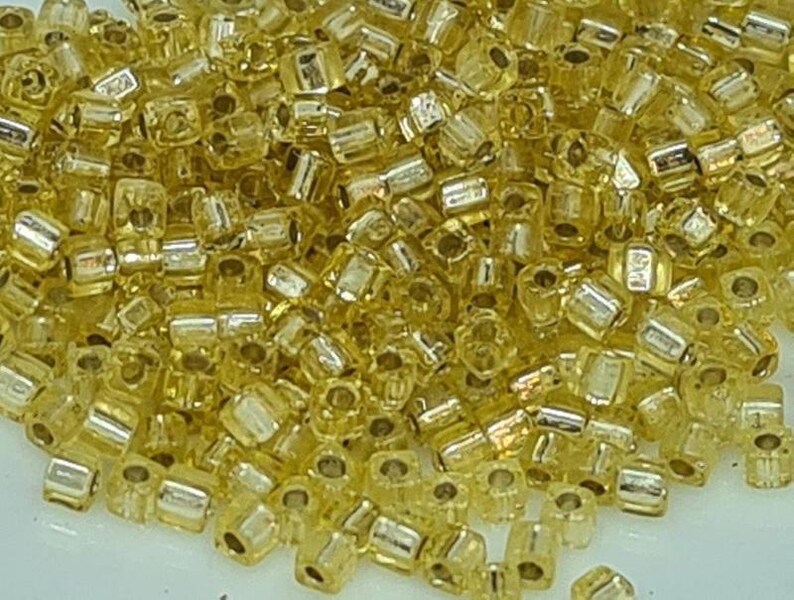 Puede incluir: Un mont&oacute;n grande de peque&ntilde;as cuentas de vidrio de color dorado. Las cuentas son redondas y tienen un acabado brillante y met&aacute;lico.