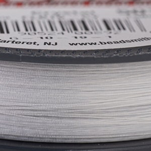 以下が含まれることがあります： 白いビーズ糸の糸巻きのクローズアップ。糸巻きにはバーコードと「Carteret, NJ www.beadsmith」のテキストが記載されたラベルが付いています。糸はきつく巻かれており、明るくニュートラルな色に見えます。