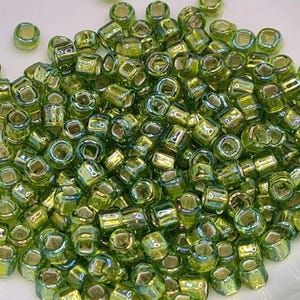 Pode incluir: Um grande monte de contas de sementes de vidro verde iridescente. As contas s&atilde;o pequenas e de forma quadrada.