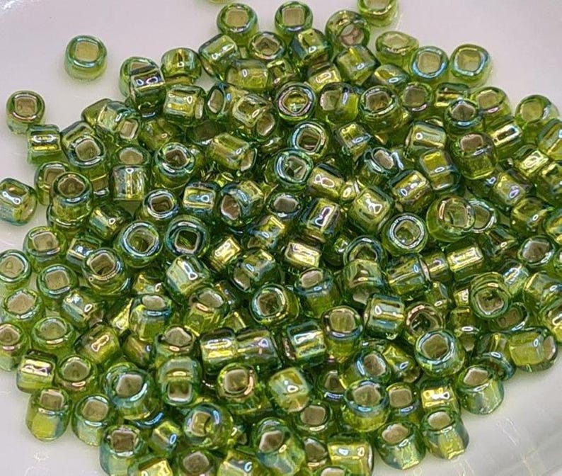 Pode incluir: Um grande monte de contas de sementes de vidro verde iridescente. As contas s&atilde;o pequenas e de forma quadrada.