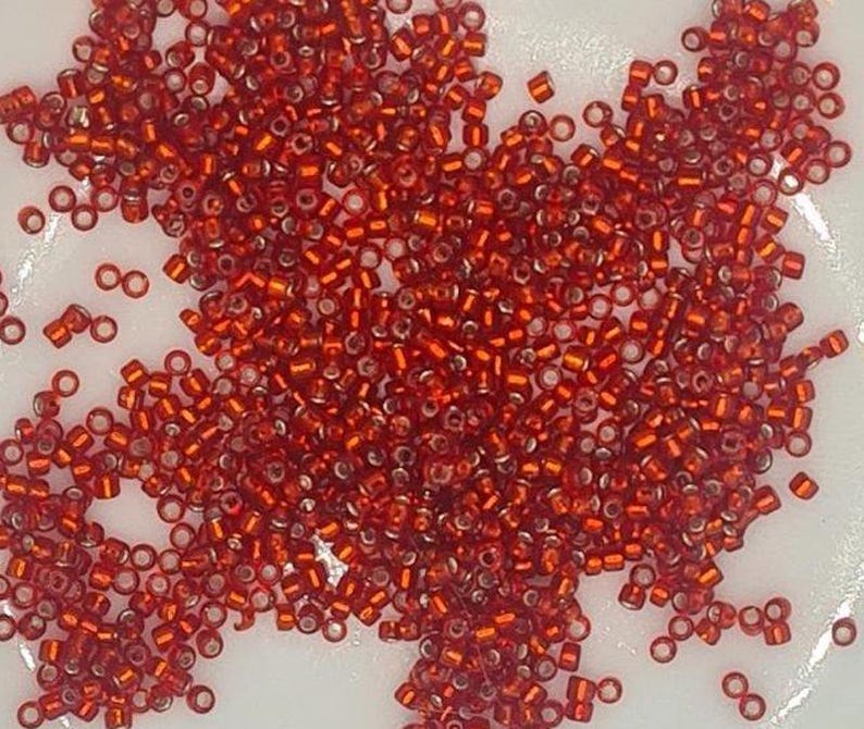 May include: A large pile of small, red, glass seed beads. The beads are translucent and have a slight shimmer.