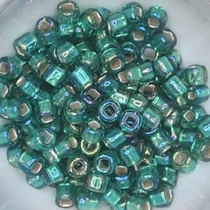 Pode incluir: Uma grande quantidade de pequenas contas quadradas de vidro de cor turquesa com brilho iridescente. As contas t&ecirc;m um furo prateado no centro.