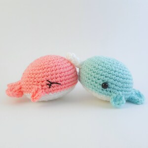 May include: Two crocheted whale plushies, one pink and one blue, are cuddling together. They have white bellies and black eyes.