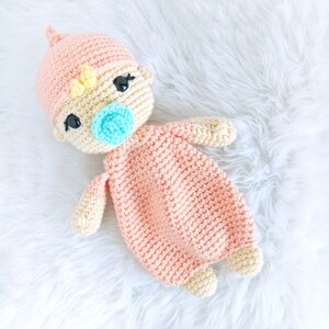 May include: A crocheted baby doll with a pink and yellow outfit and a blue pacifier. The doll is lying on a white fluffy surface.
