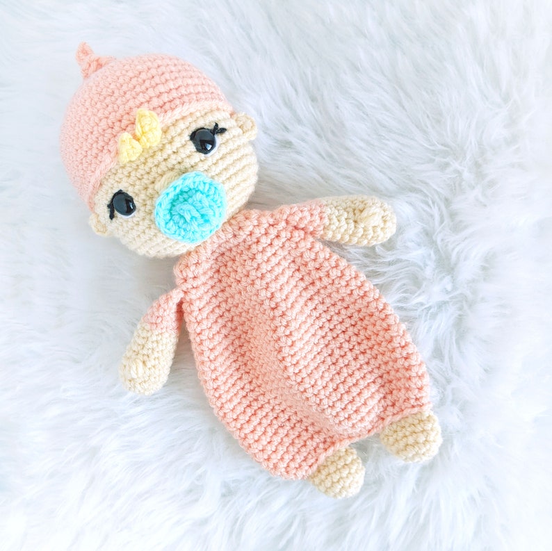 May include: A crocheted baby doll with a pink and yellow outfit and a blue pacifier. The doll is lying on a white fluffy surface.