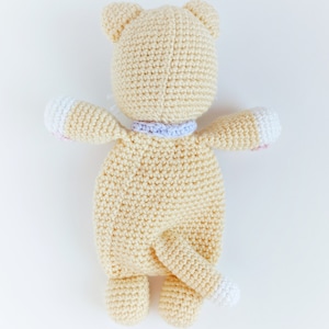 May include: A crocheted stuffed animal in the shape of a cat. The cat is cream colored with a white tail and pink accents on the paws. The cat is wearing a purple and white collar.