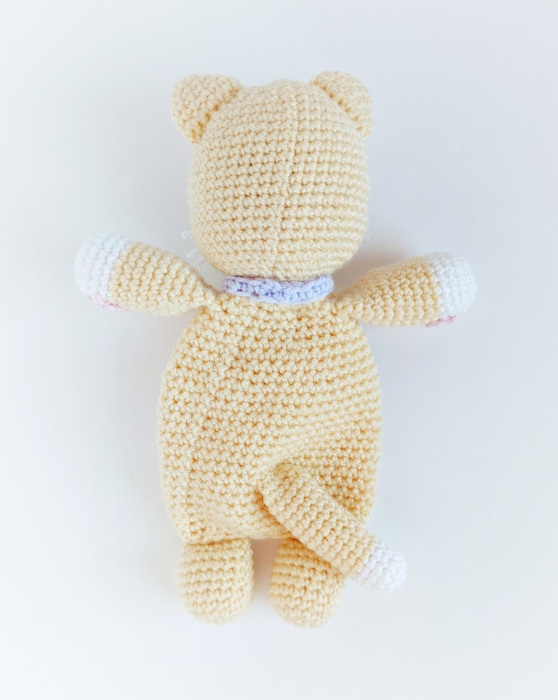 May include: A crocheted stuffed animal in the shape of a cat. The cat is cream colored with a white tail and pink accents on the paws. The cat is wearing a purple and white collar.