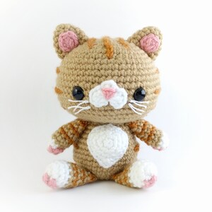 May include: A crocheted orange tabby cat with white accents. The cat has pink ears and a white belly. The cat is sitting on a white background.