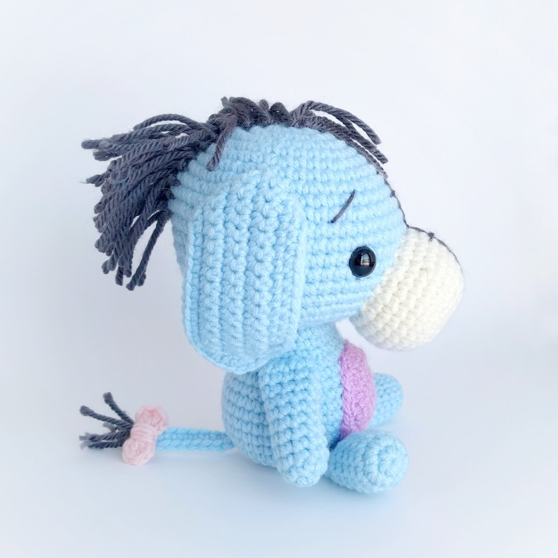 May include: A blue crocheted stuffed animal resembling a donkey with a gray mane, white snout, and black eyes. The donkey has a pink tail and is sitting on a white surface.