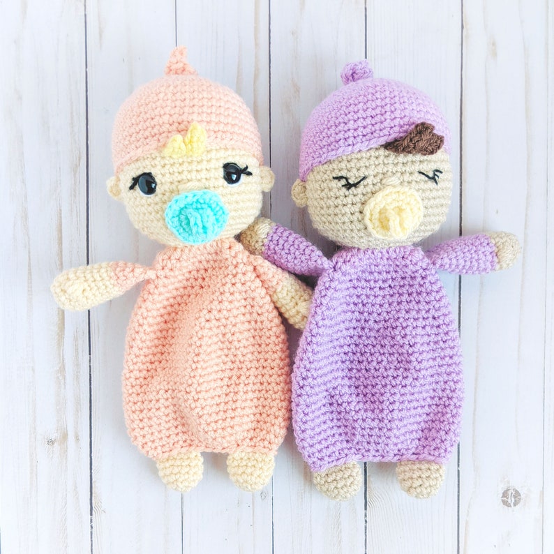 May include: Two crocheted baby dolls, one in peach and one in lavender, with embroidered facial features and pacifiers. The dolls are lying on a white wooden surface.