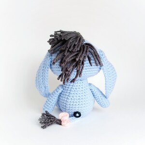 May include: A blue crocheted stuffed animal with a brown yarn mane and tail. The animal has a button nose and is sitting on a white background.