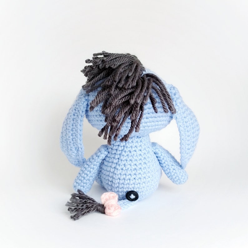May include: A blue crocheted stuffed animal with a brown yarn mane and tail. The animal has a button nose and is sitting on a white background.