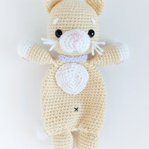 May include: A crocheted cream-colored cat toy with white accents. The cat has black eyes, a pink nose, and a white heart on its chest.