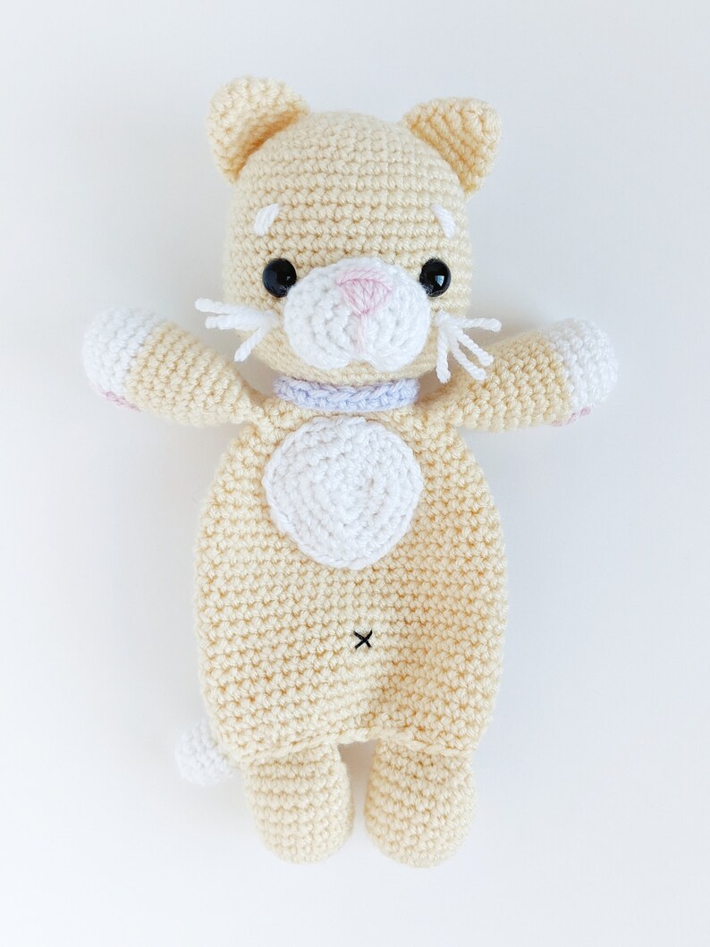 May include: A crocheted cream-colored cat toy with white accents. The cat has black eyes, a pink nose, and a white heart on its chest.