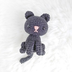 May include: A small crocheted black cat with pink nose and black eyes. The cat is sitting on a white fluffy surface.