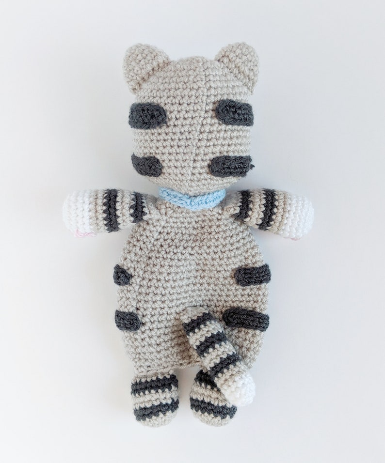 May include: A crocheted gray and black striped cat toy with a blue collar. The cat has a white belly and a white tip on its tail.