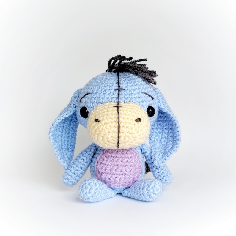 May include: A crocheted stuffed animal donkey with blue fur, a yellow nose, and black eyes. The donkey is sitting on a white surface.