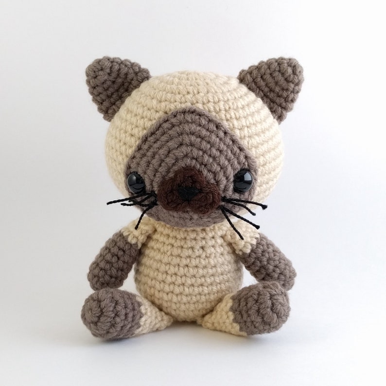 May include: A crocheted Siamese cat toy with brown and cream colored fur, black eyes, and a black nose. The cat is sitting with its legs crossed.
