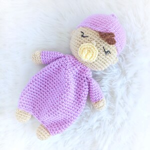 May include: A crocheted baby doll with a purple outfit and a pink hat. The doll has a pacifier and brown embroidered eyebrows.