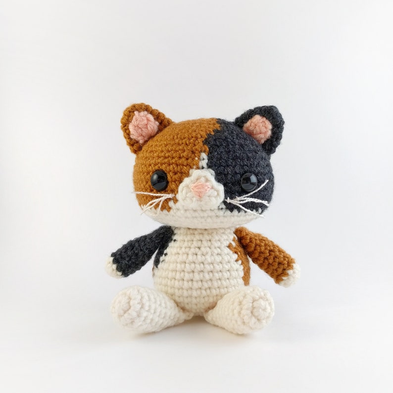 May include: A crocheted calico cat toy with black, white, and orange fur. The cat has black eyes and a pink nose.