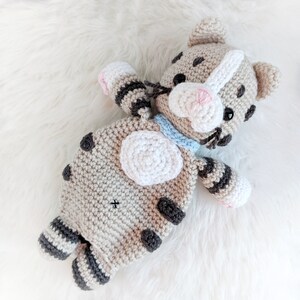 May include: A crocheted stuffed animal cat toy with brown and white stripes, a white belly, and a light blue collar. The cat has a pink nose and black eyes.