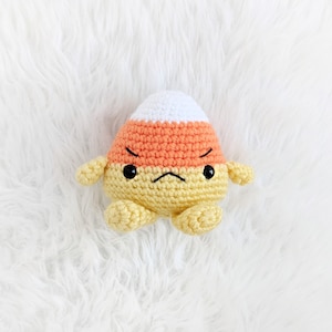 May include: A crocheted candy corn toy with a grumpy expression. The toy is orange, yellow, and white.
