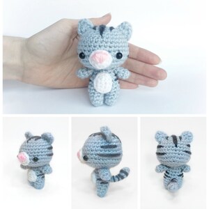 May include: A small gray and black striped crocheted cat toy with white accents. The cat is standing on a white surface.