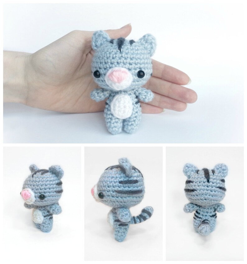 May include: A small gray and black striped crocheted cat toy with white accents. The cat is standing on a white surface.